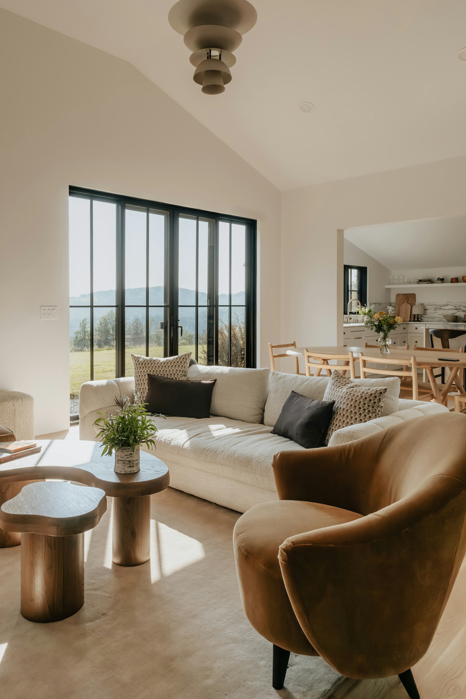 A sunlit living room with a cream sofa, a brown armchair, wooden tables, and large windows overlooking wooded hills